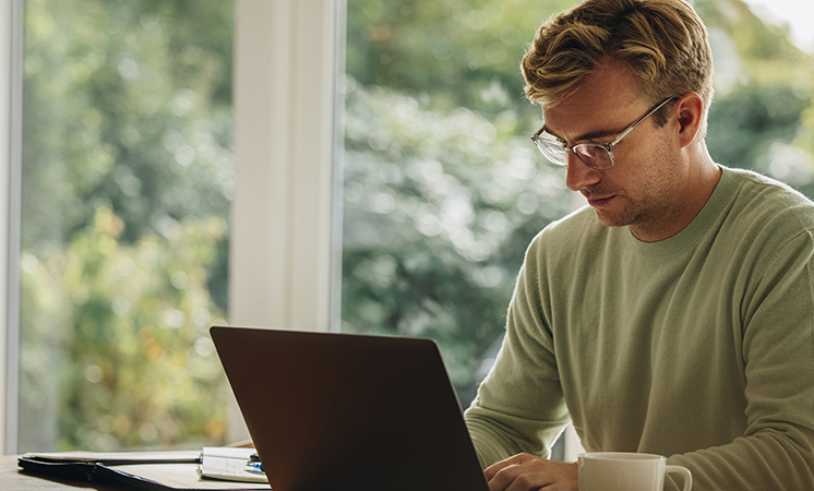junger Mann mit Brille arbeitet konzentriert an seinem Laptop. Neben ihm steht eine Kaffeetasse.